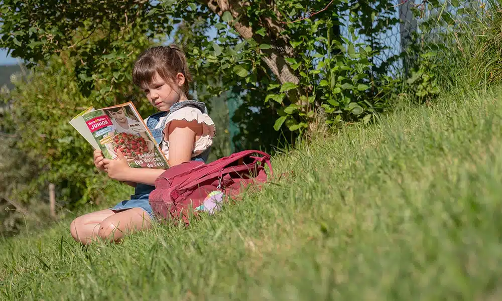 Image d'une jeune fille assise sur une pente herbeuse, sous un arbre, lisant le magazine Terre&Nature Junior.