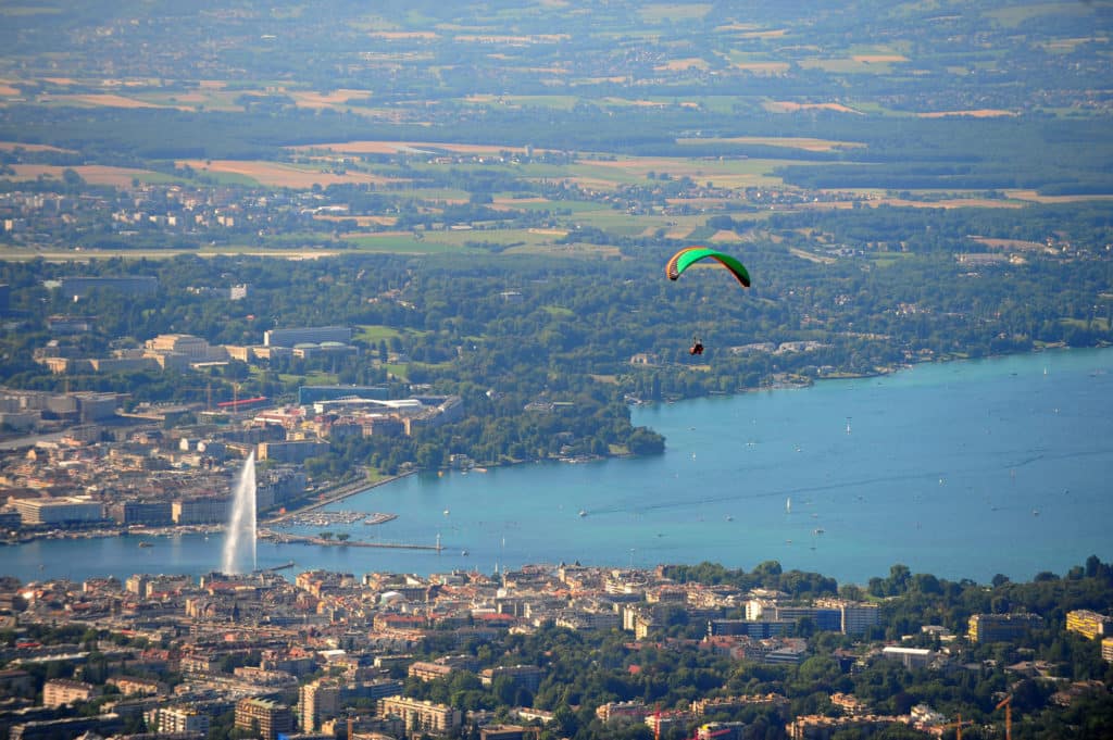 Parapentiste avec voile verte survolant un lac et une ville avec jet d'eau, paysage urbain et campagne en arrière-plan.