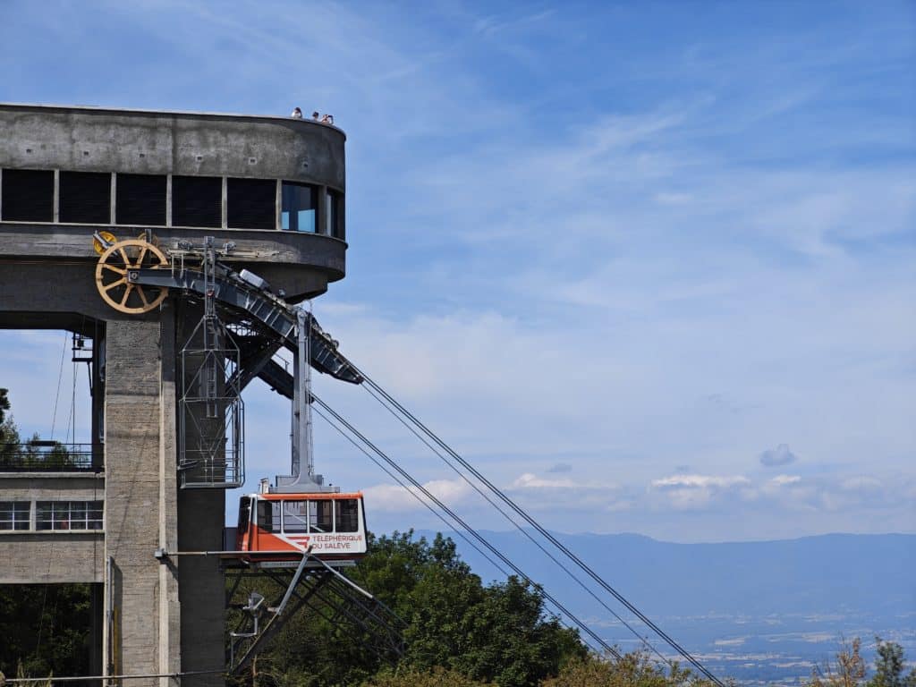 Station supérieure de téléphérique en béton avec plateforme d'observation, cabine orange et grande poulie, dominant une vue panoramique sur les montagnes.