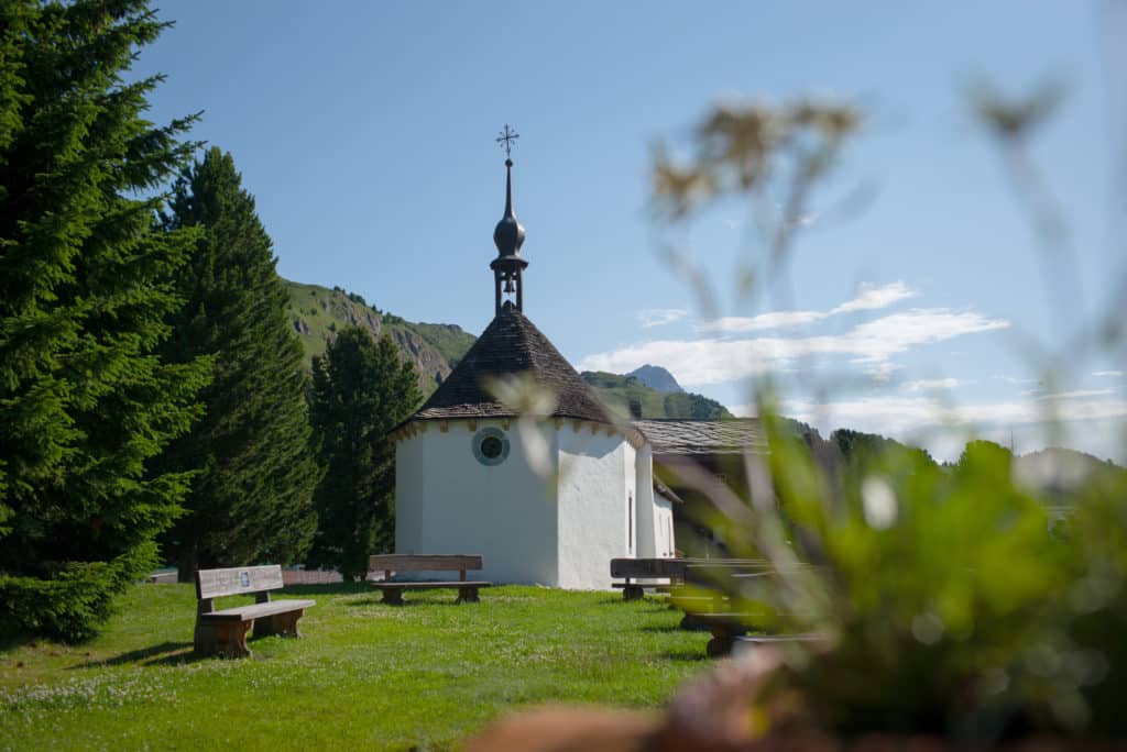 Petite chapelle alpine blanche avec clocher à bulbe dans une prairie verdoyante entourée de sapins, avec montagnes en arrière-plan et fleurs au premier plan.