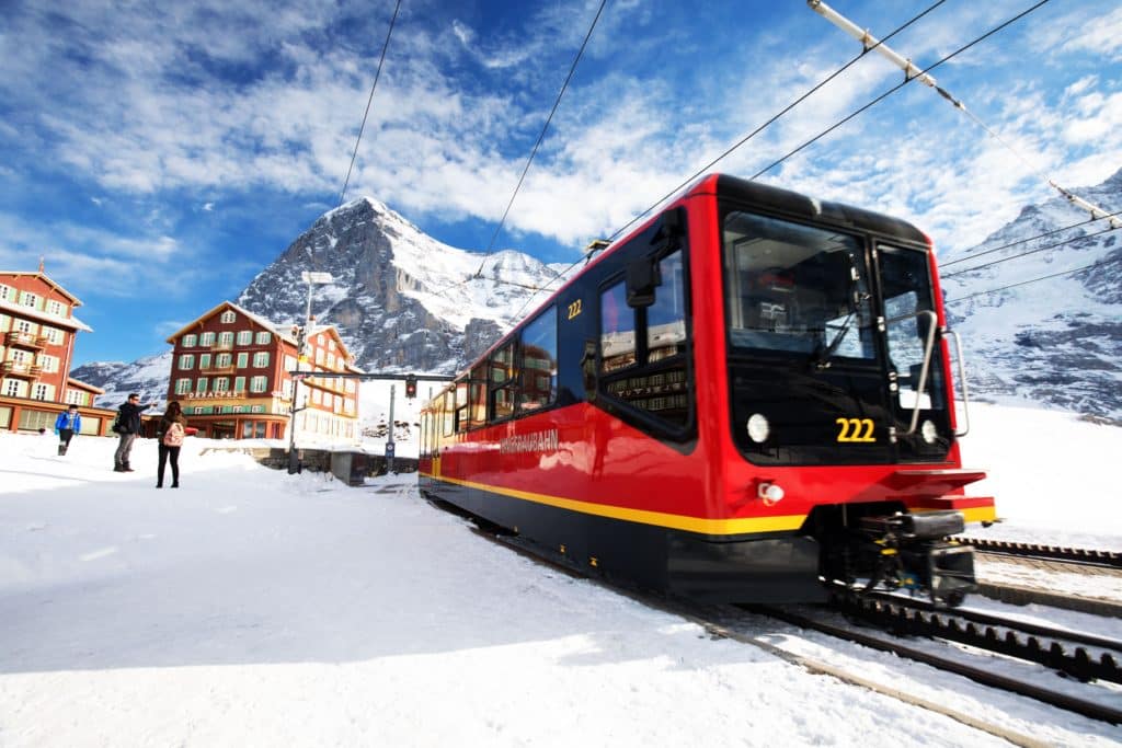 Train à crémaillère rouge et noir en gare de montagne enneigée avec chalets traditionnels et sommet alpin en arrière-plan sous un ciel bleu nuageux.