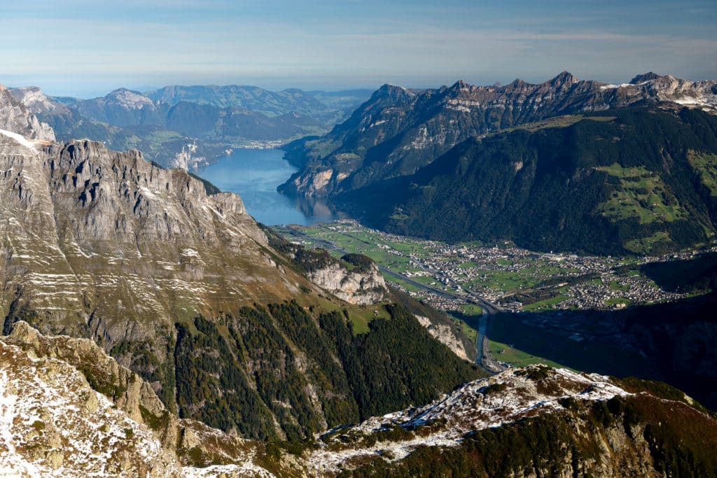 Vue panoramique alpine depuis un sommet montrant un lac entouré de montagnes, avec villages et prairies dans la vallée en contrebas.