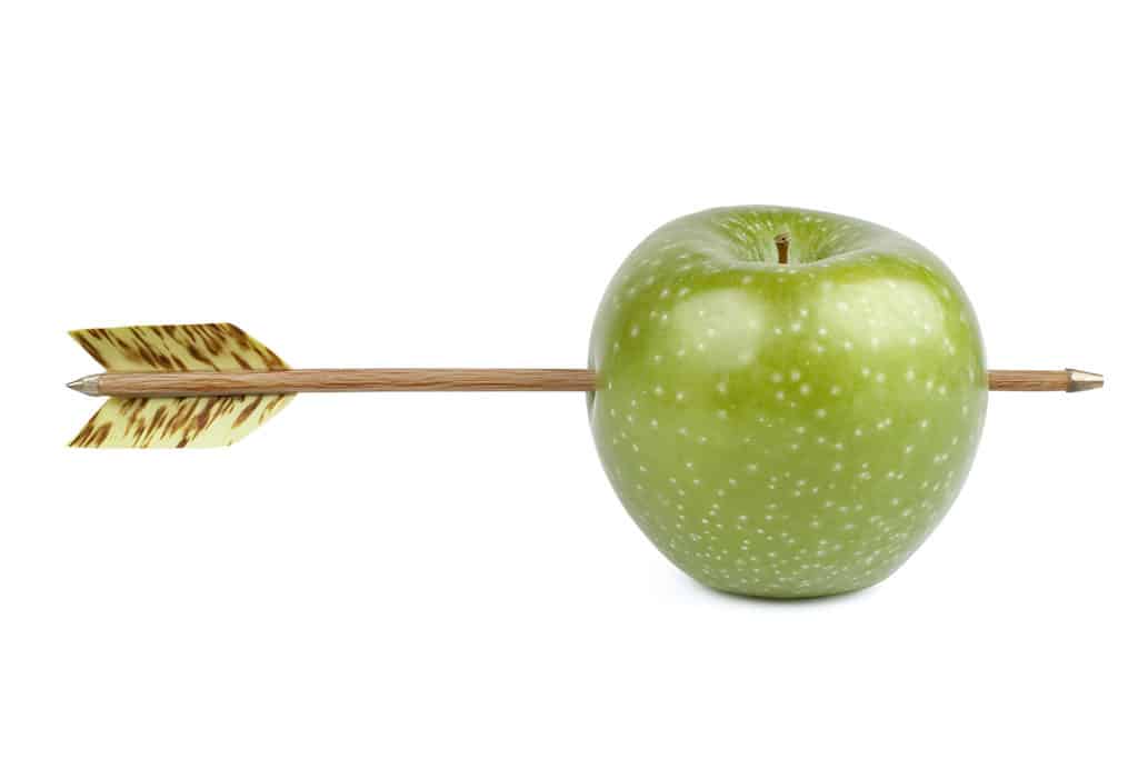 Pomme verte traversée par une flèche en bois avec empennage, sur fond blanc.