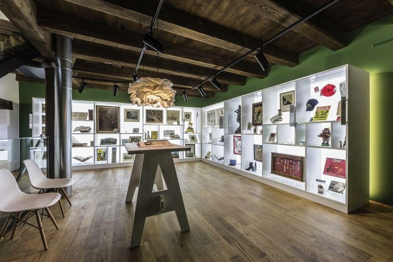 Salle d'exposition avec murs verts, plafond en bois apparent, étagères murales blanches présentant diverses œuvres d'art et objets, table centrale en bois et chaises design modernes.