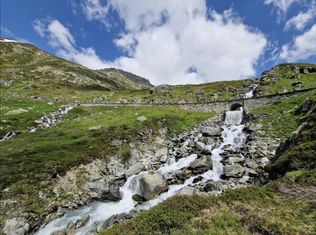 Torrent de montagne dévalant un alpage verdoyant avec pont en pierre et cascade, sous un ciel bleu nuageux.