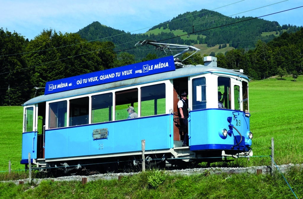 Tramway historique bleu clair avec publicité Le Média traversant une prairie verdoyante avec montagnes boisées en arrière-plan.